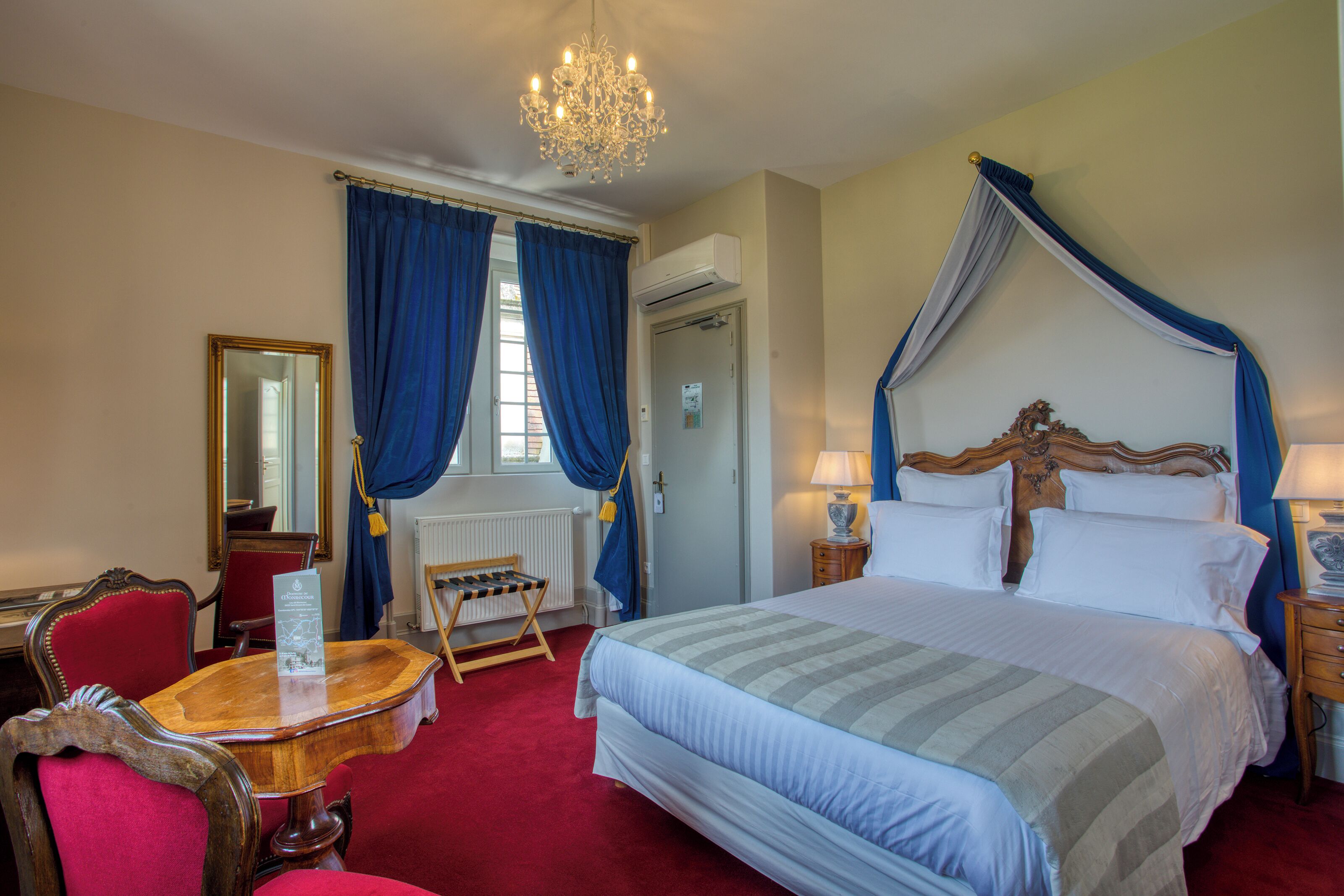 A hotel room with a four-poster bed, a wooden chair, a small table, and a chandelier.