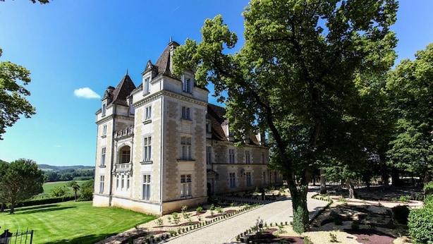 A large, historic stone building with multiple stories and a prominent tower, surrounded by well-maintained gardens and a clear blue sky. - Domaine du Château de Monrecour – Hôtel et Restaurant (Saint-Vincent-de-Cosse)