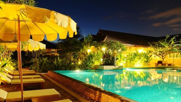 Une piscine entourée de plantes tropicales, agrémentée de chaises longues et de parasols, et bordée d’un bâtiment dont le toit est recouvert de tuiles.