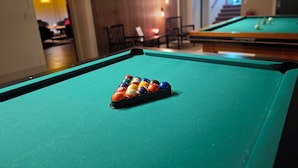 Una mesa de billar con un portaobjetos lleno de bolas y una sala con paneles de madera y una escalera al fondo.
