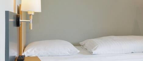 Quarto de hotel com cama, almofadas brancas, controle remoto e luminária fixada na parede.