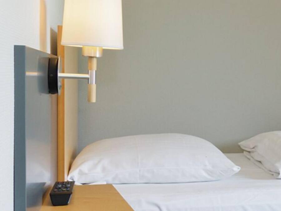 Une chambre d’hôtel avec un lit, des oreillers blancs, une télécommande et une lampe fixée au mur.