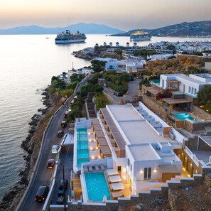 A coastal hotel with a pool, a road adjacent to the sea, and cruise ships in the background.