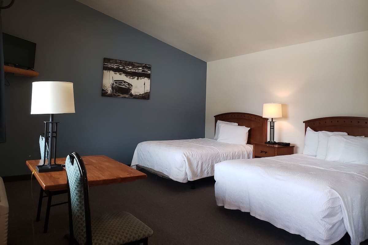 A hotel room with two beds, a desk, a chair, a lamp, and a painting on the wall.