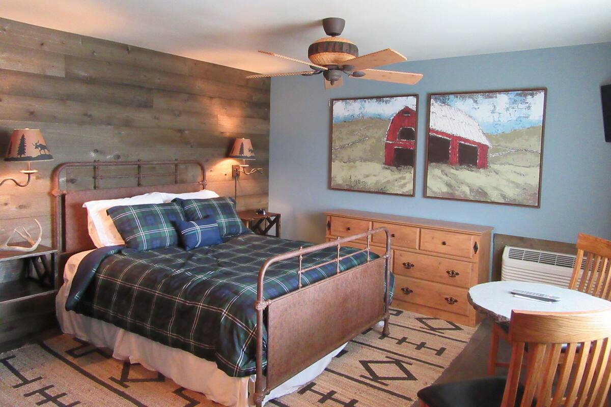 A bedroom with a wooden headboard, a bed with plaid bedding, a ceiling fan, two wall-mounted lamps, a dresser, a chair, and two paintings of barns.