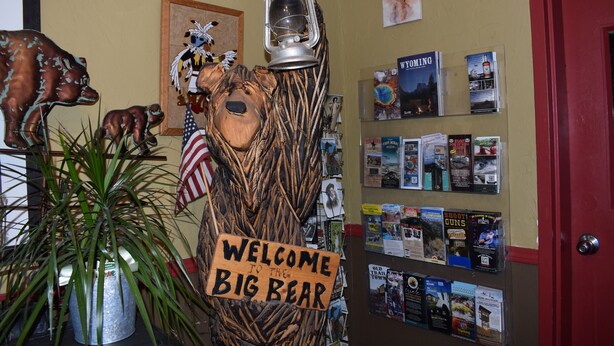 Un letrero de madera que dice 'Welcome to the Big Bear', con una figura de oso y un farol.
