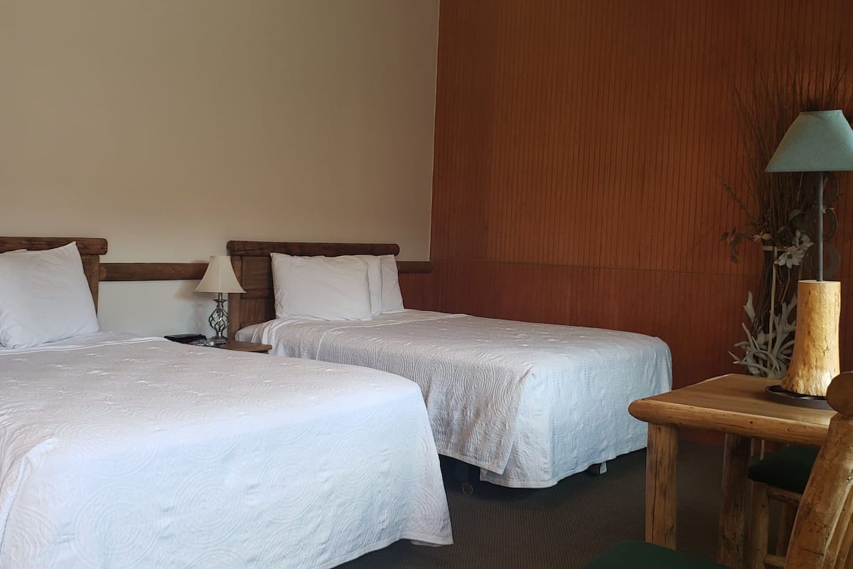 A hotel room with two beds, a wooden nightstand, a table with a lamp, and a chair.
