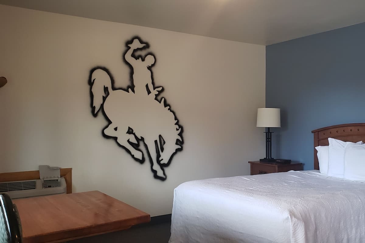 A hotel room with a large wall art of a rooster, a bed with white bedding, a wooden nightstand with a lamp, and a small table with a chair.