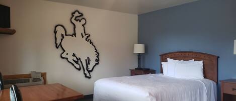 A hotel room with a large wall art of a rooster, a bed with white bedding, a wooden nightstand with a lamp, and a small table with a chair.