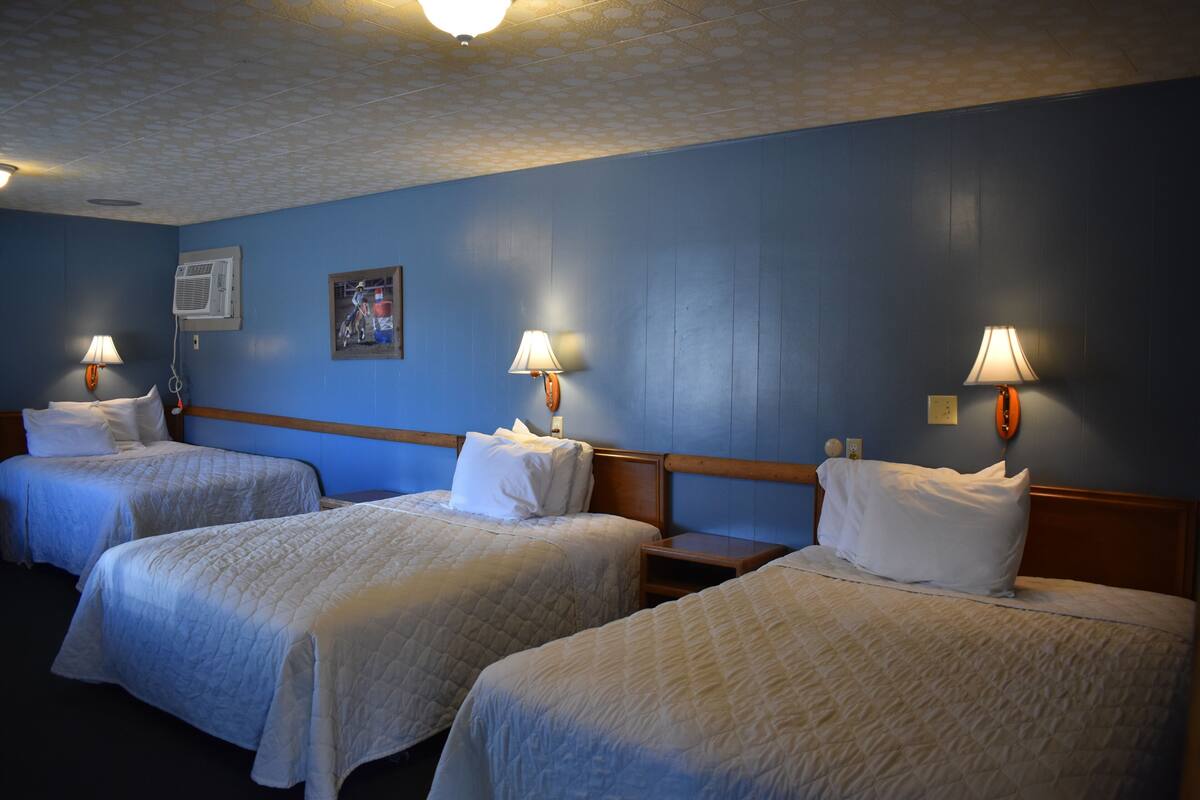 A hotel room with three beds, each with a white bedspread and pillows. There are wall-mounted lamps on either side of the beds.