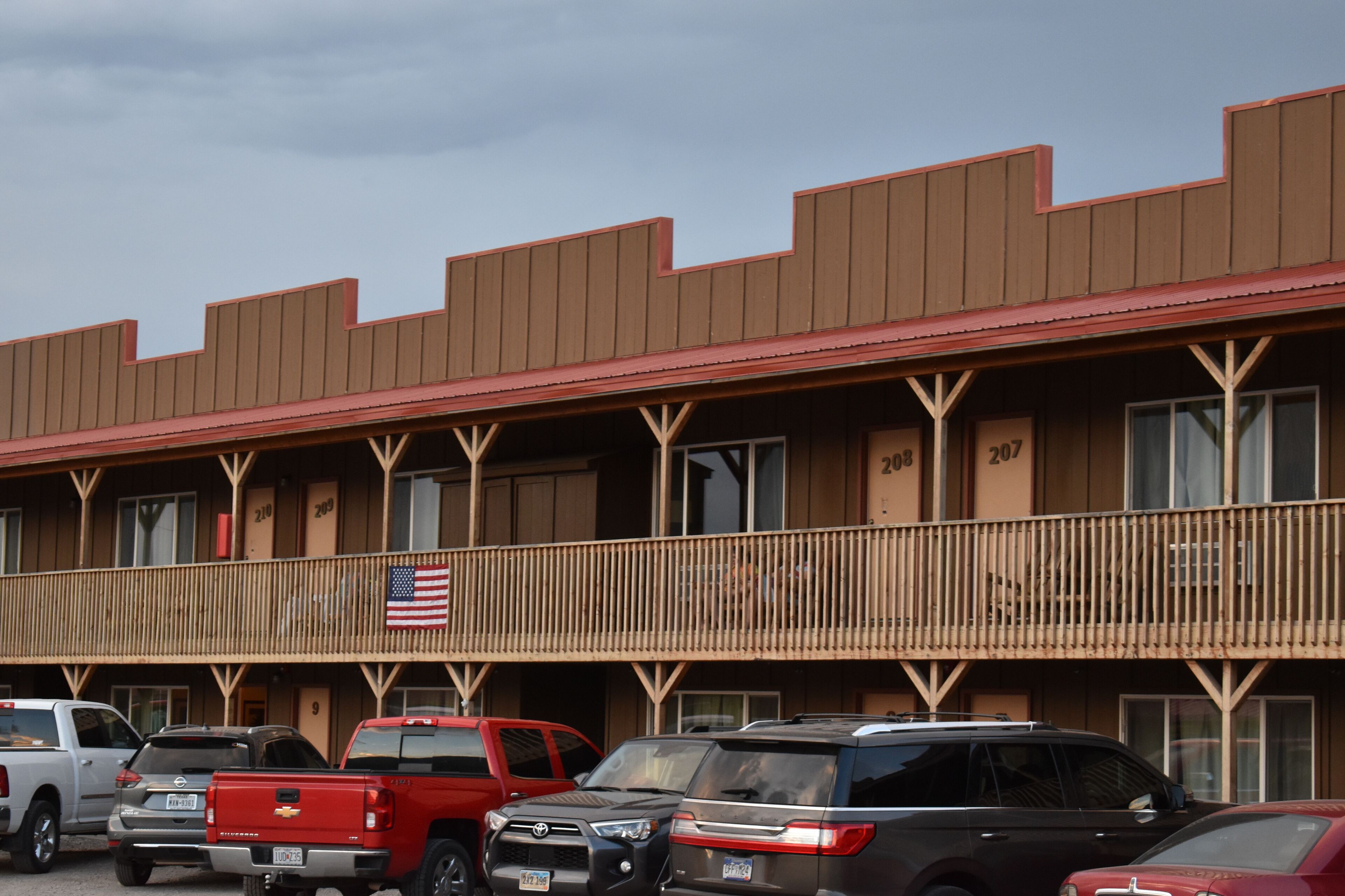A motel with a red roof and a balcony with an American flag. There are several parked vehicles in front.