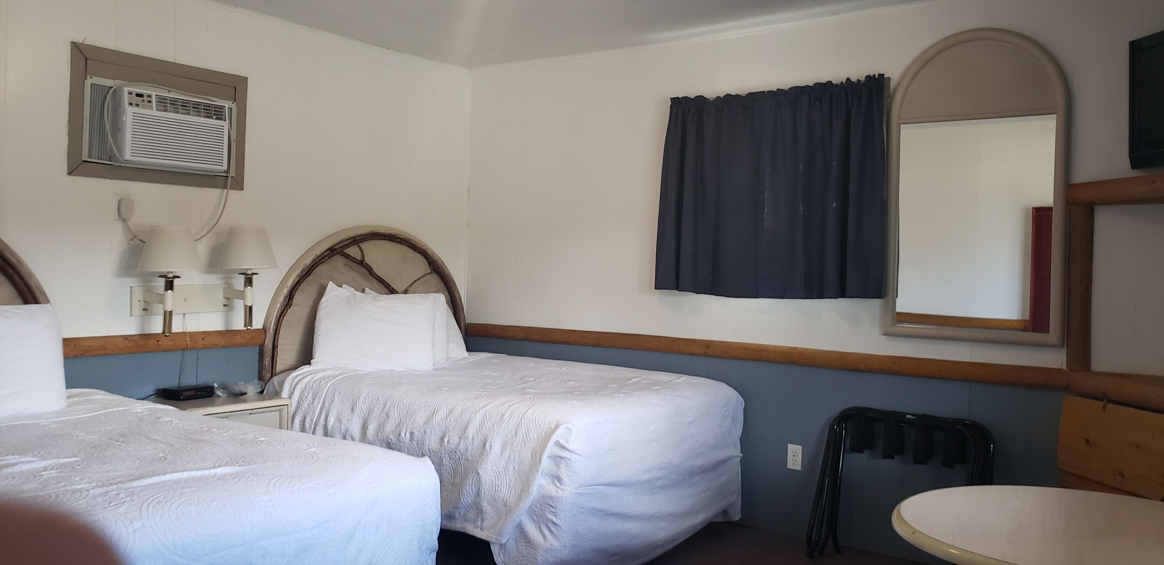 A hotel room with two beds, a small table, a chair, a mirror, a window with curtains, and an air conditioning unit.