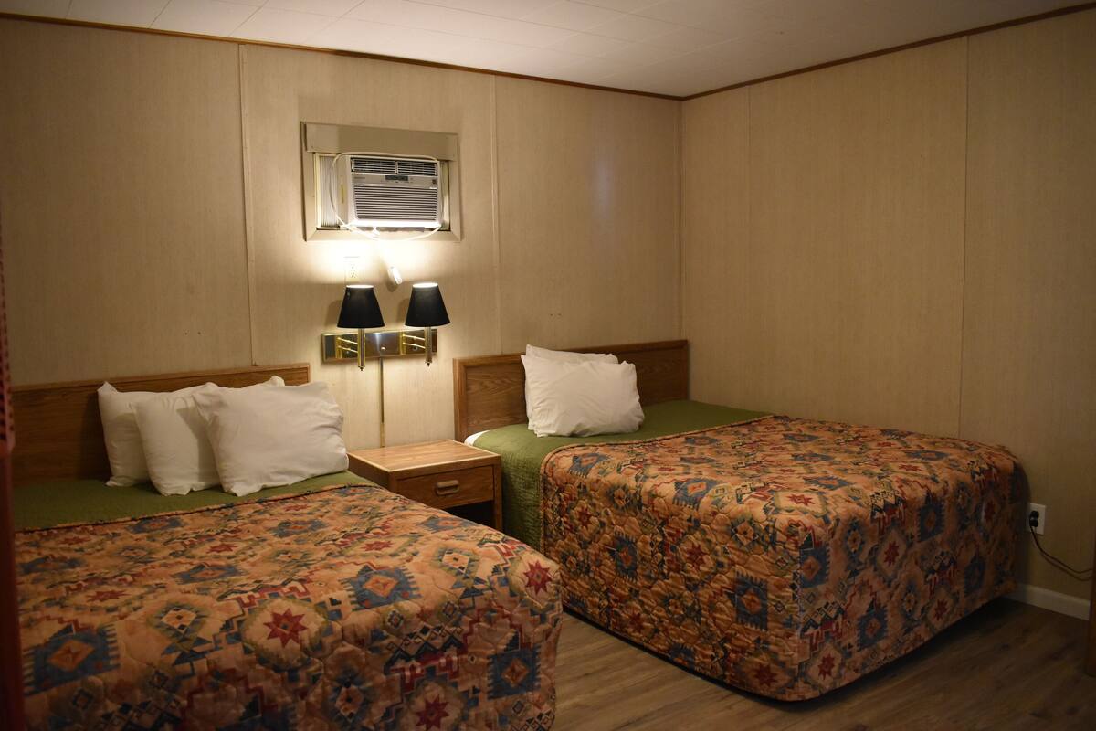 A hotel room with two beds, each with a patterned bedspread, a nightstand with two lamps, and a window with blinds.
