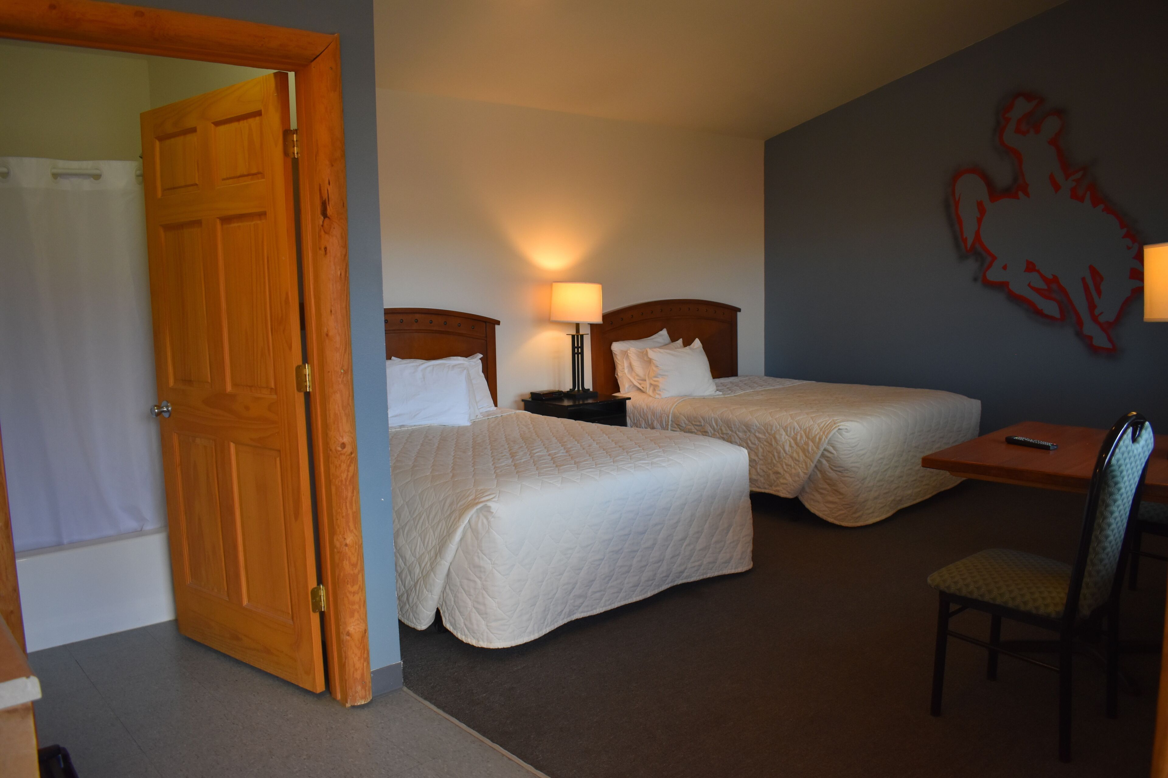 A hotel room with two beds, a desk, a chair, a lamp, and a wooden door.