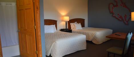 A hotel room with two beds, a desk, a chair, a lamp, and a wooden door.