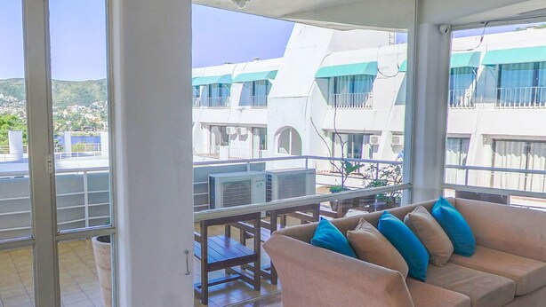 Un balcon avec un canapé beige, une table et des chaises. De grandes fenêtres offrent une vue sur des immeubles et une étendue d’eau.