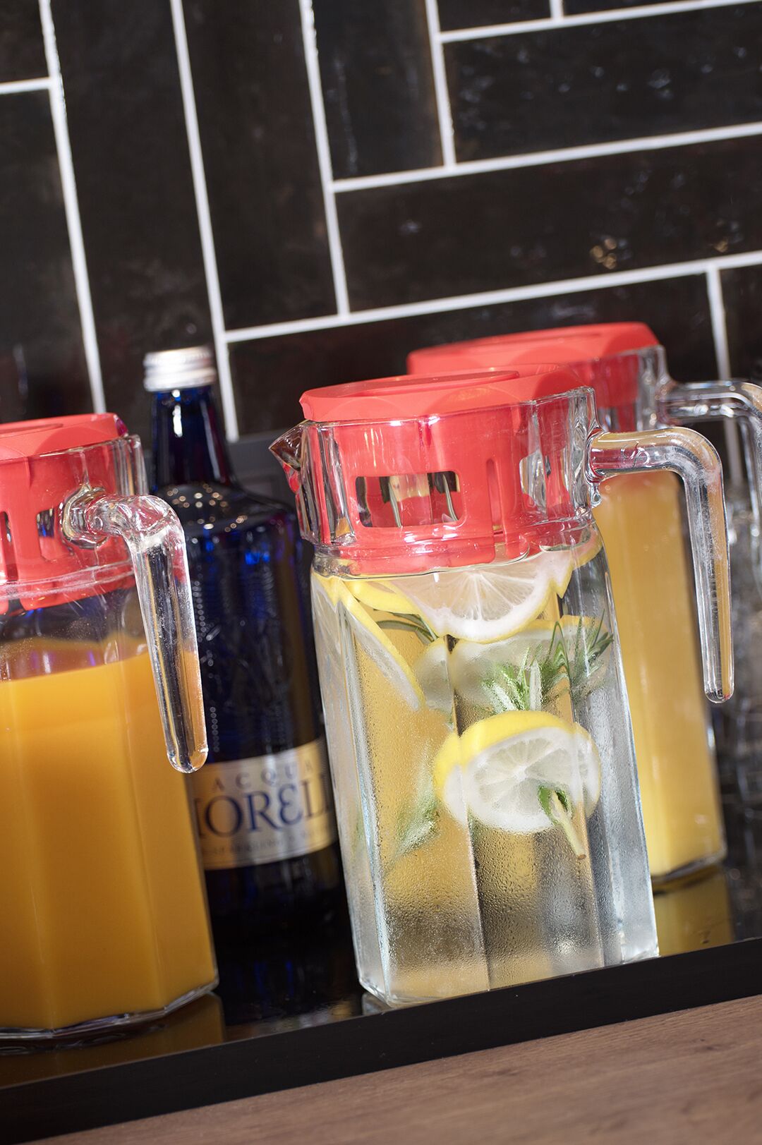 Un comptoir de cuisine sur lequel reposent trois carafes en verre remplies de boissons, dont une avec un couvercle rouge et les deux autres avec des couvercles transparents.
