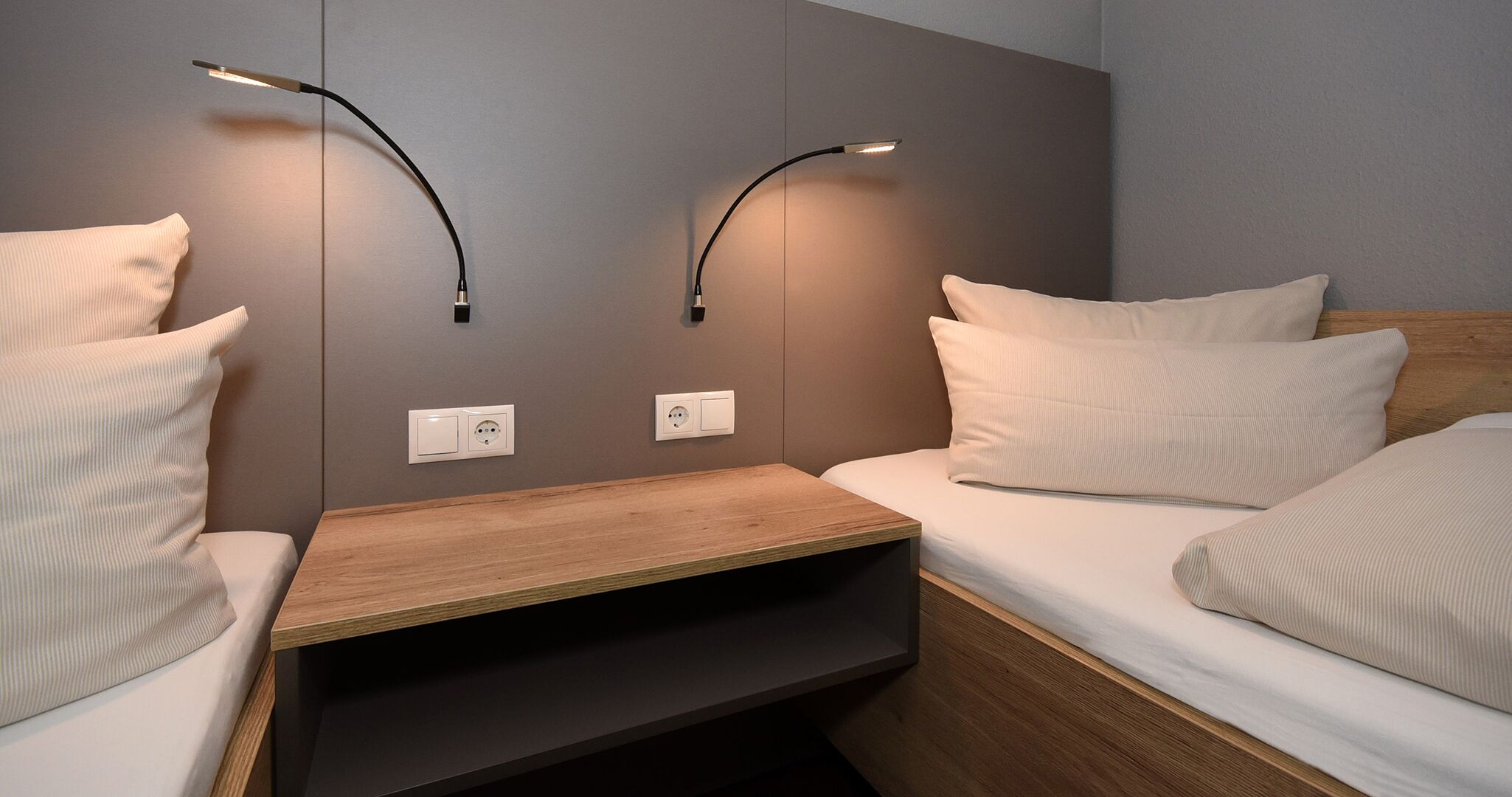 Ein modernes Hotelzimmer mit einem Holfnachtschränkchen, zwei Nachttischen und einem Bett mit weißer Bettwäsche. An der Wand befinden sich zwei Wandleuchten zum Lesen und zwei Steckdosen.