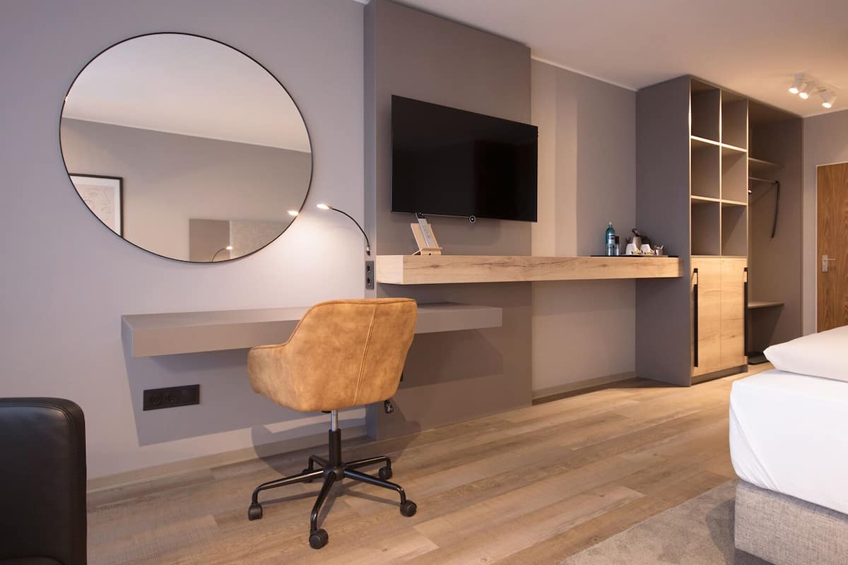 Habitació d'hotel moderna amb escriptori de fusta, mirall rodó, televisió, llit i armari.
