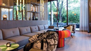 A modern living room with a sectional sofa, a colorful ottoman, and a glass coffee table. There is a bookshelf with decorative items and a large window offering a view of the outdoors. - Motel One Wien-Prater (Vienna)