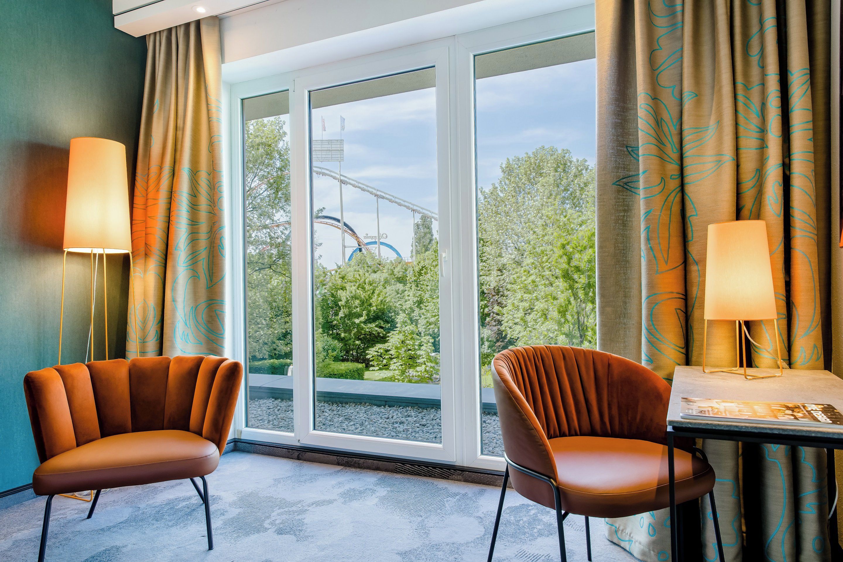 Ein Hotelzimmer mit zwei Sesseln, einem Tisch und Blick auf eine Achterbahn.