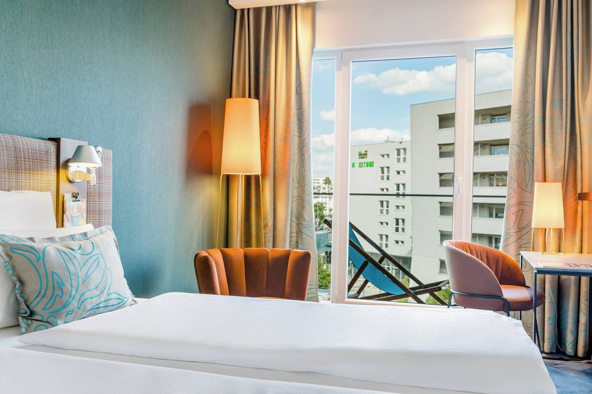 A hotel room with a bed, two chairs, a table, and a view of buildings through a large window.