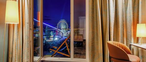 A hotel room with a view of a Ferris wheel at night, a chair, a table, and curtains.