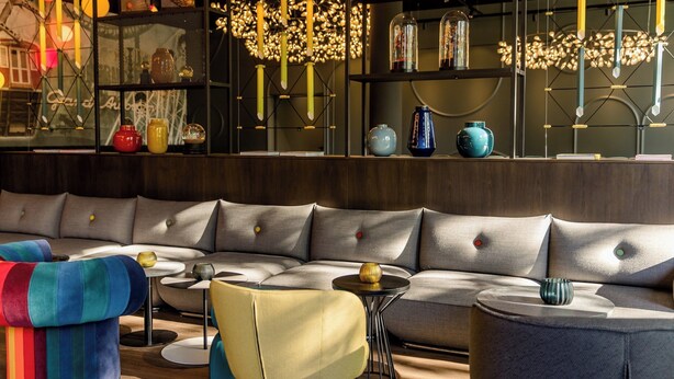 A modern interior with a sectional sofa, colorful chairs, a small table, and decorative items on shelves. - Motel One Wien-Prater (Vienna)