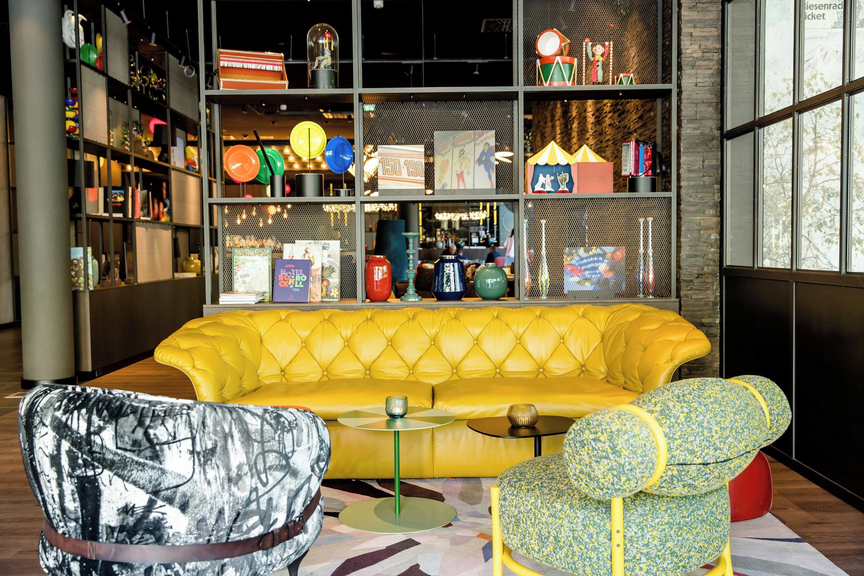 A vibrant room with a yellow sofa, a patterned armchair, and a small table. Shelves in the background display various items including books, decorative objects, and a sign with text.