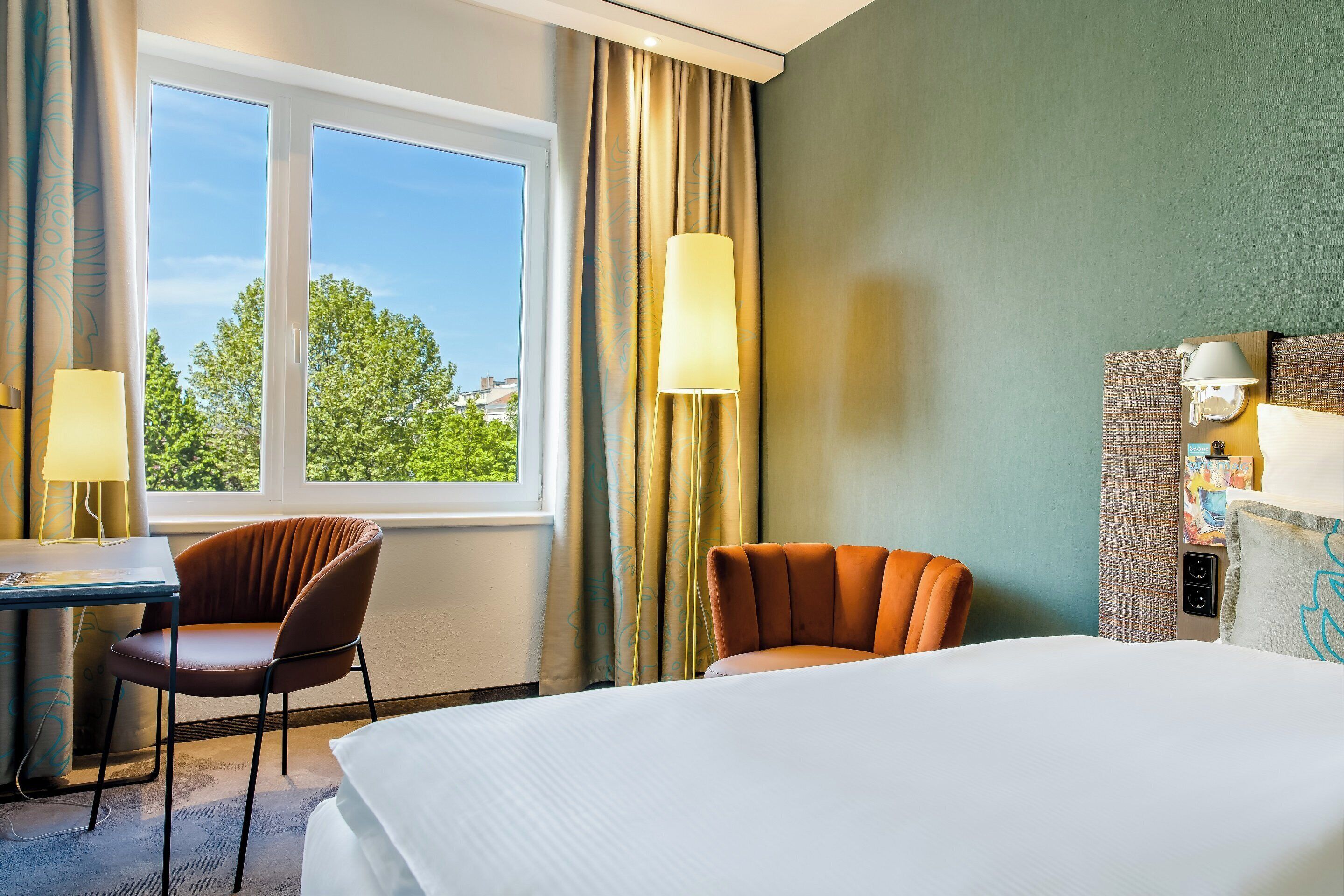 A hotel room with a bed, two chairs, a desk, a lamp, and a view of trees through the window.