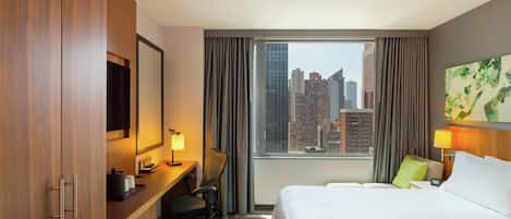 Habitación de hotel con cama, escritorio, silla y un ventanal con vistas a la ciudad.