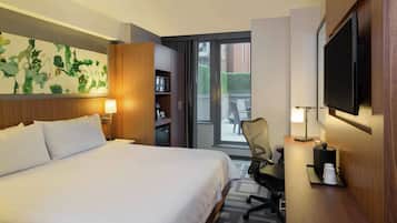 A hotel room with a large bed, a desk with a chair, a flat-screen TV, a bookshelf, and a sliding door to a balcony.