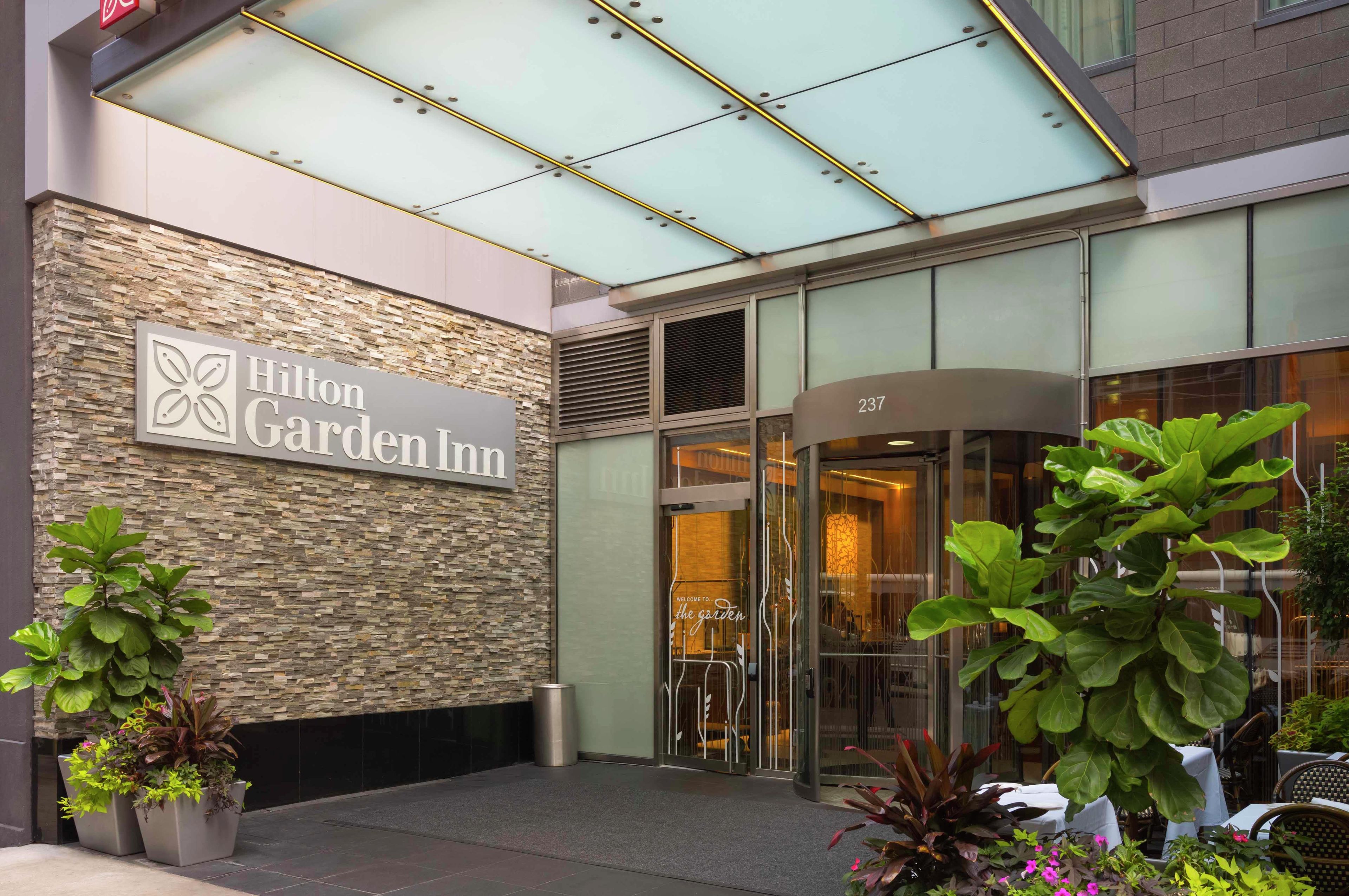 L’extérieur d’un hôtel Hilton Garden Inn, avec une façade en pierre, une entrée en verre et des plantes en pot.
