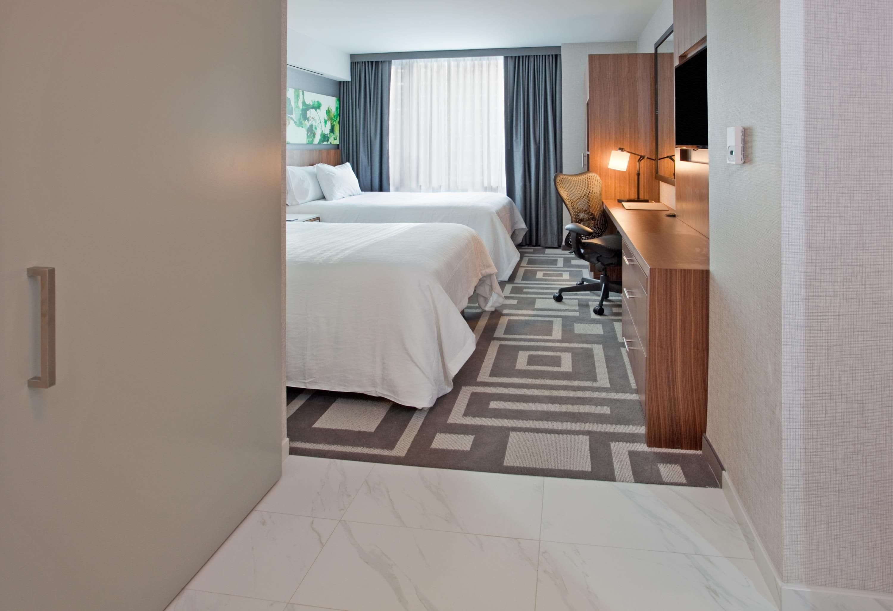 A hotel room with two beds, a desk, a chair, a TV, and a window with curtains.