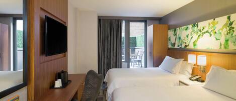 A hotel room with a bed, a desk with a TV, a chair, a bedside table with a lamp, and a large window with curtains.