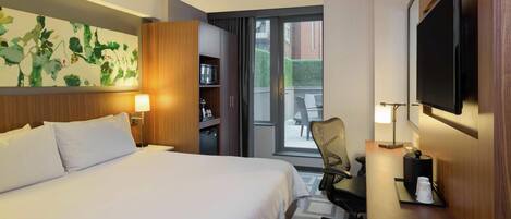 A hotel room with a bed, a desk with a chair, a television, a bookshelf, and a sliding door to a balcony.