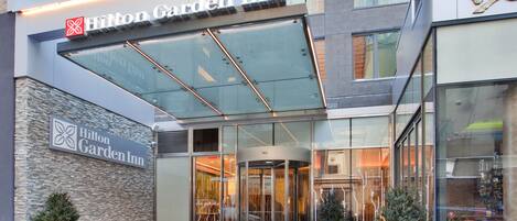 Exterior de um Hilton Garden Inn, com entrada de vidro, fachada de pedra e plantas em vasos.