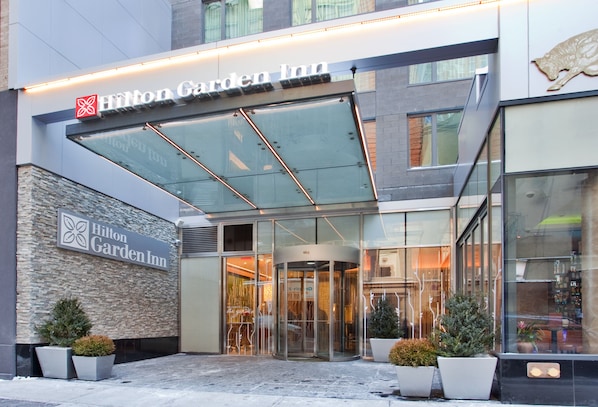 Exterior de um Hilton Garden Inn, com entrada de vidro, fachada de pedra e plantas em vasos.