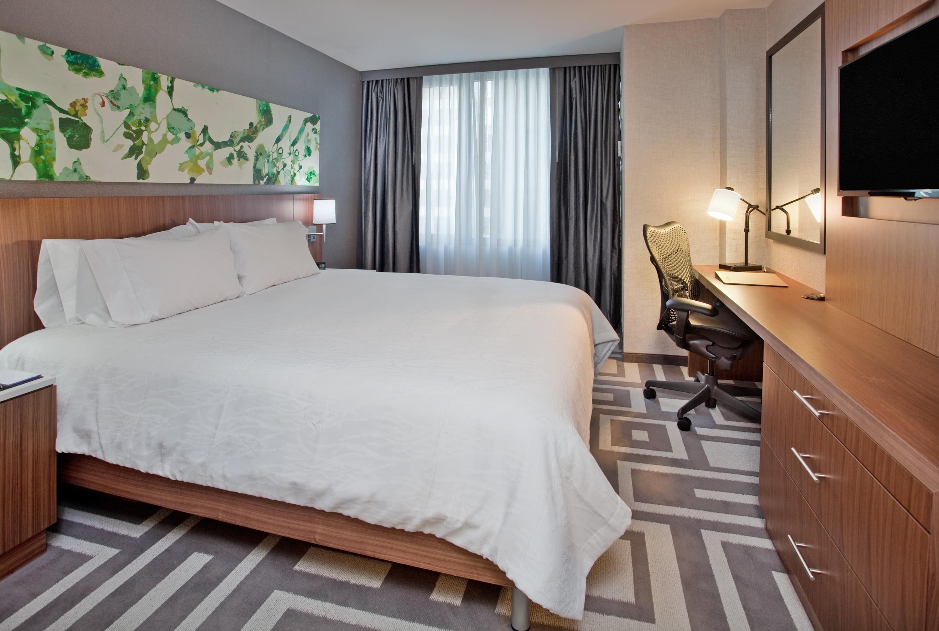 A hotel room with a bed, a desk with a chair, a television, and a large window with curtains.