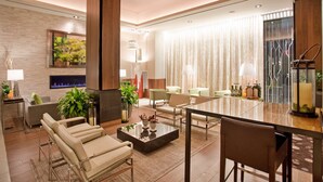 A modern hotel lobby with a fireplace, comfortable seating, a dining area, and a large window with curtains. - Hilton Garden Inn New York/Central Park South-Midtown West (New York)