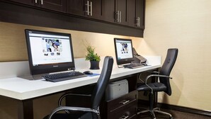 A modern office space with two desks, each with a laptop and a desktop monitor. There are two black office chairs, a small potted plant, and a printer on the desks. The room has dark wooden cabinets above the desks and a beige carpet. - Hampton Inn Los Angeles Int'l Airport/Hawthorne (Hawthorne)