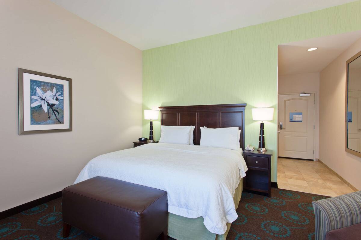 A hotel room with a large bed, a nightstand, a lamp, a bench, and a door leading to another room.