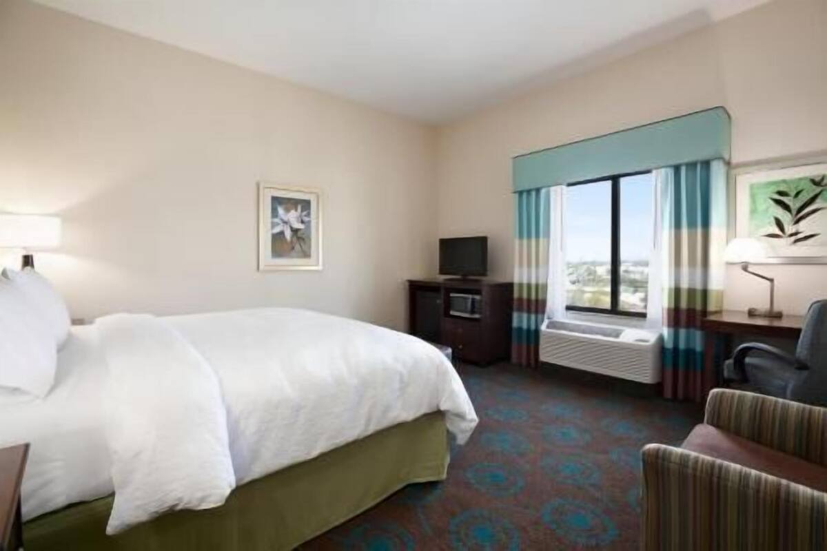 A hotel room with a bed, a desk with a computer, a TV, a window with curtains, and a chair.