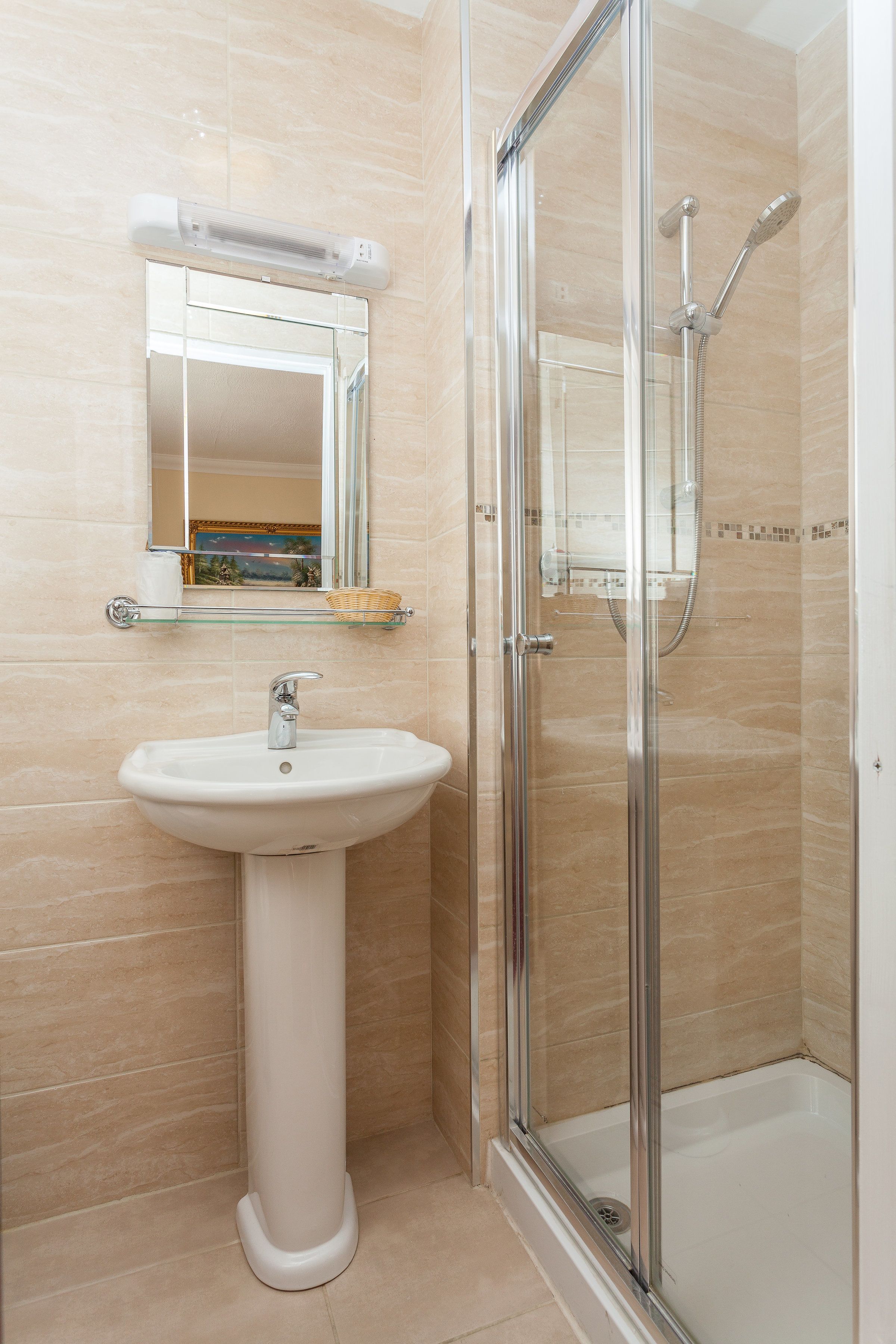A bathroom with a glass shower enclosure, a pedestal sink, and a mirror with a shelf above it.