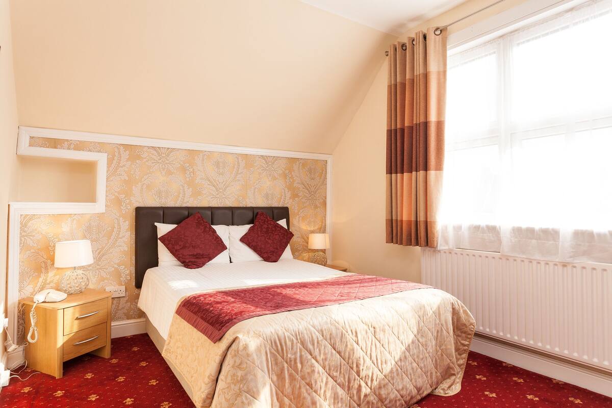 A hotel room with a bed, a nightstand with a phone, a window with curtains, and a radiator.