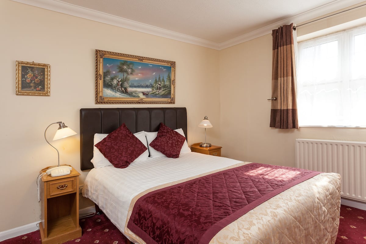 A neatly arranged bedroom with a large bed, two bedside tables, a nightstand, a lamp, a framed painting, and a window with curtains.