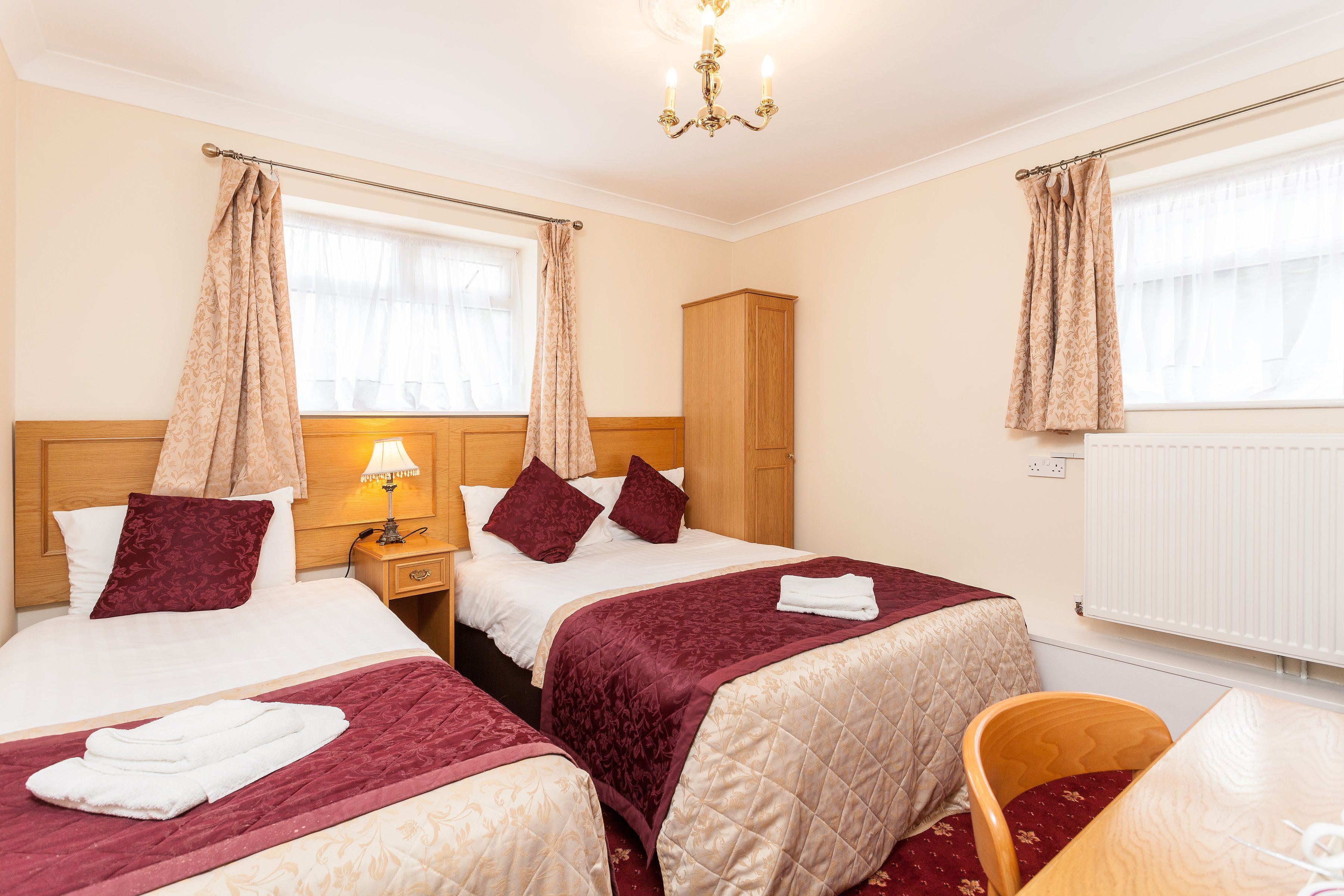 A hotel room with two beds, a wooden wardrobe, a nightstand with a lamp, a chair, and a table.