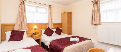 A hotel room with two beds, a wooden wardrobe, a nightstand with a lamp, a chair, and a table.