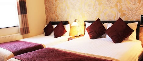 Camera d'albergo con due letti, ognuno con copripiumino e cuscini rossi, un comodino con una lampada e carta da parati decorata.