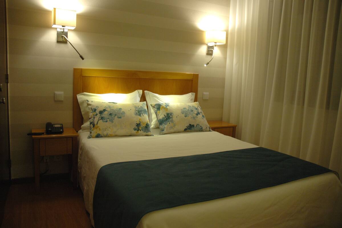 Una habitación de hotel con cama, dos lámparas de noche, una mesita de noche y una ventana con cortinas.